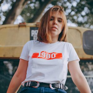 Stylish young woman in a 1980 shirt poses confidently in front of a vintage bus outdoors.